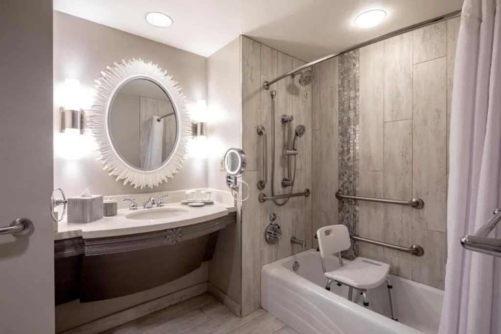 Ba&ntilde;o accesible con barras de apoyo, asiento de ba&ntilde;era y espejo redondo en Loews Royal Pacific Resort