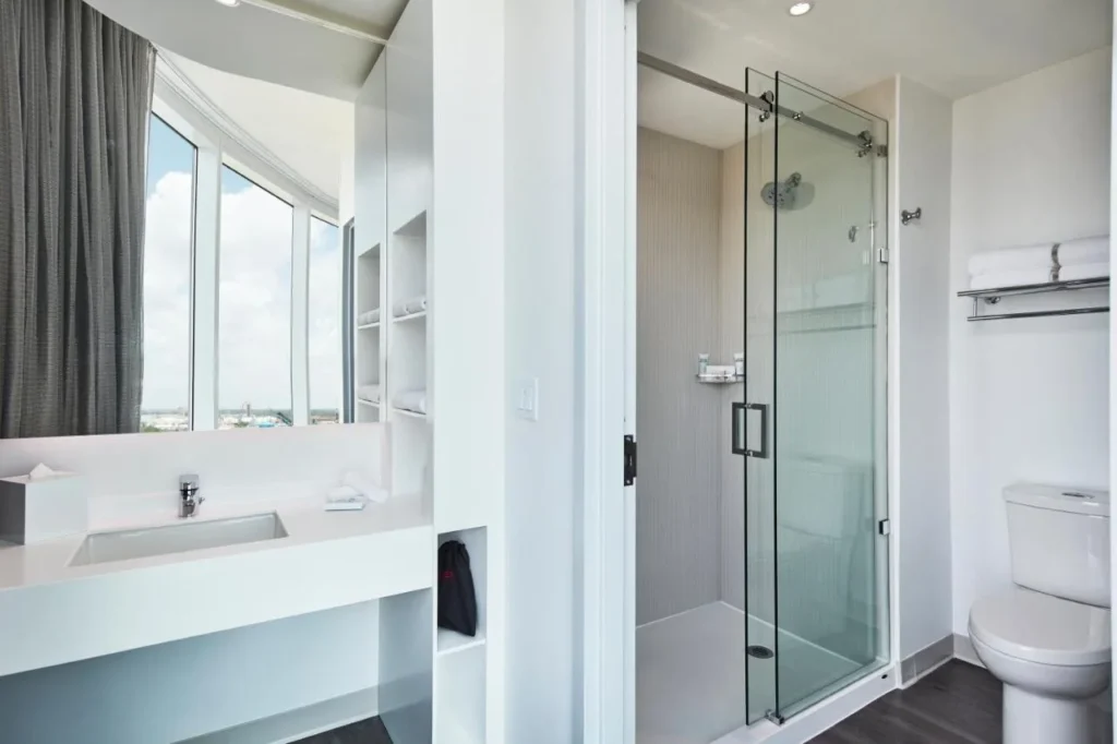 Cuarto de ba&ntilde;o con puerta corredera de ducha de cristal, tocador blanco y estantes para toallas en Universal Aventura Hotel