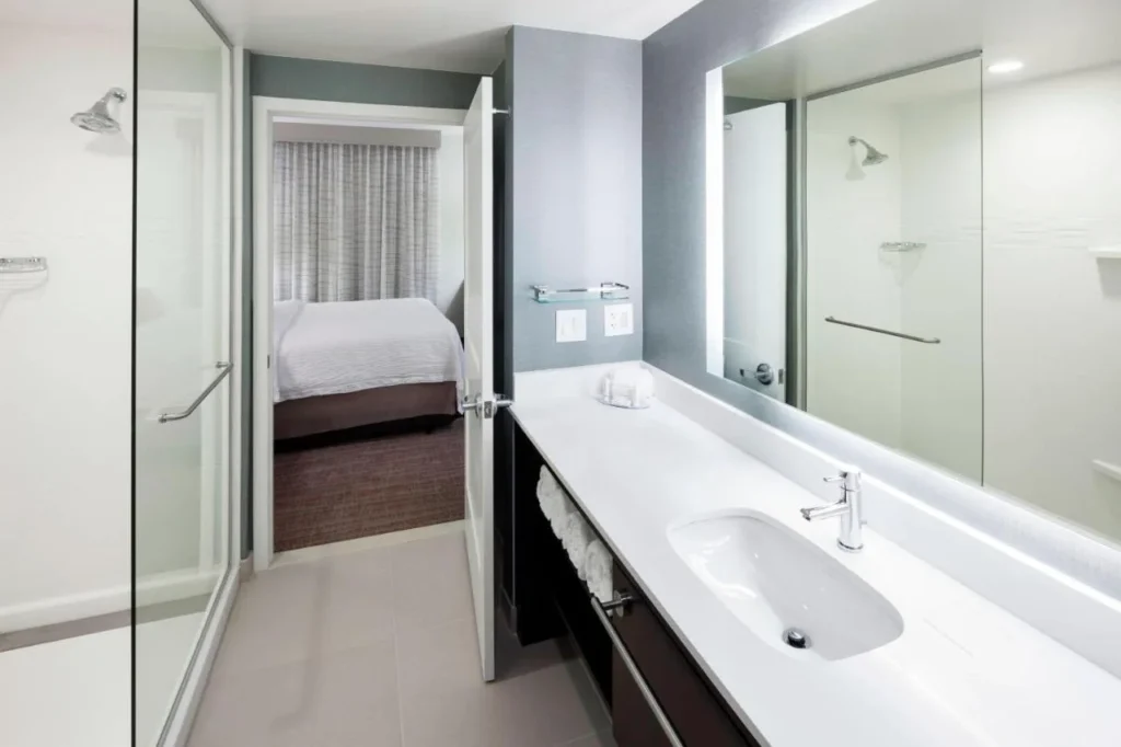 Cuarto de ba&ntilde;o con ducha a ras de suelo y tocador, dormitorio m&aacute;s all&aacute;, en el Residence Inn Near Universal Orlando