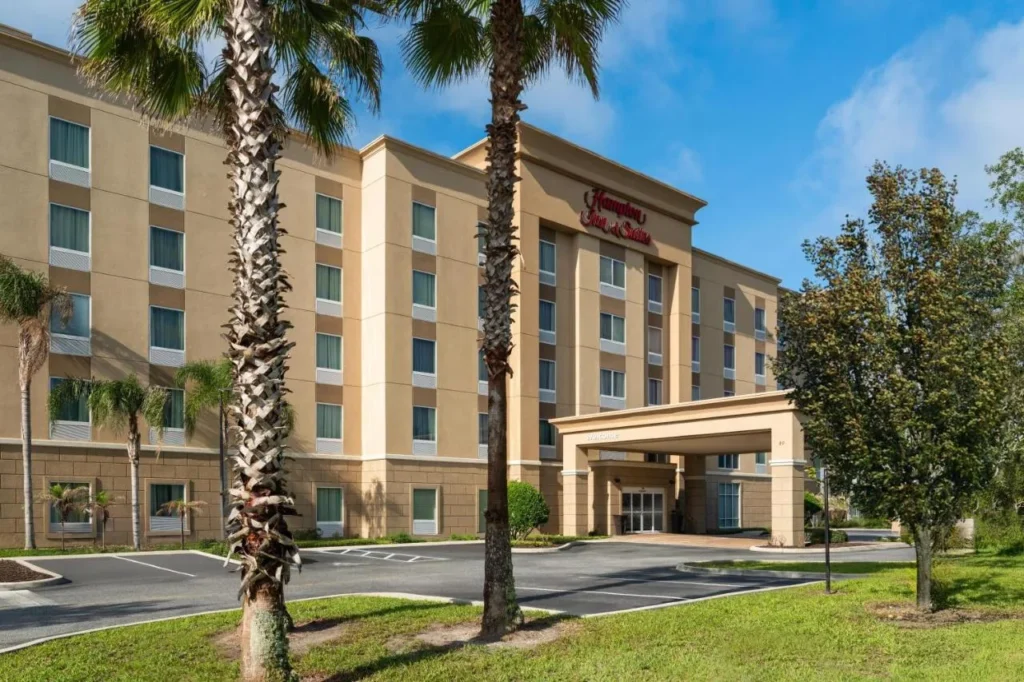 Beige multi-story exterior and entrance canopy of Hampton Inn and Suites DeLand behind palm trees