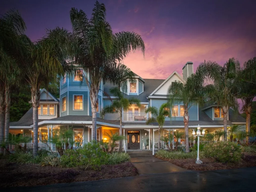 Blue Victorian mansion lit at dusk behind tall palms at Heron Cay Lakeview Bed & Breakfast