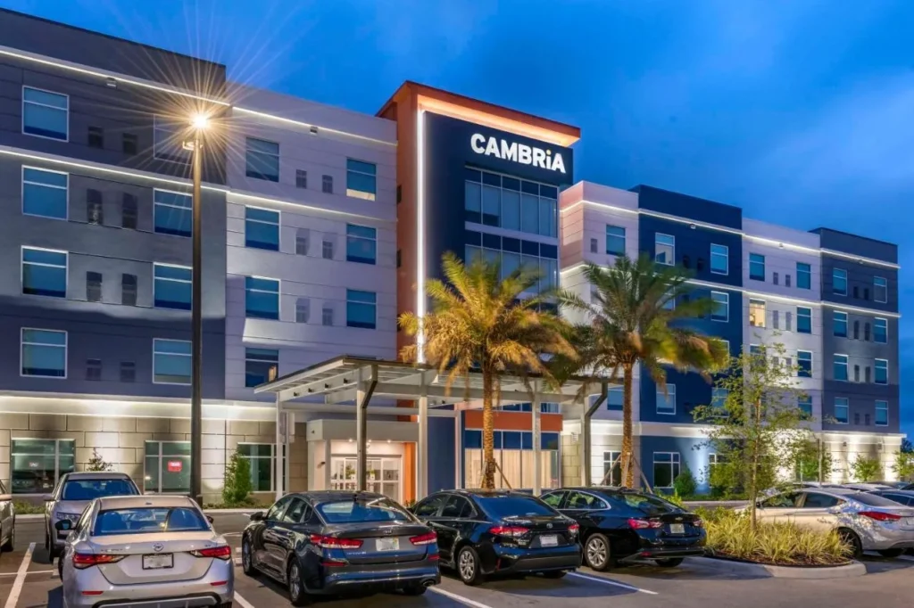 Cambria Hotel Orlando Aeropuerto fachada con letrero iluminado, palmeras y coches estacionados al atardecer