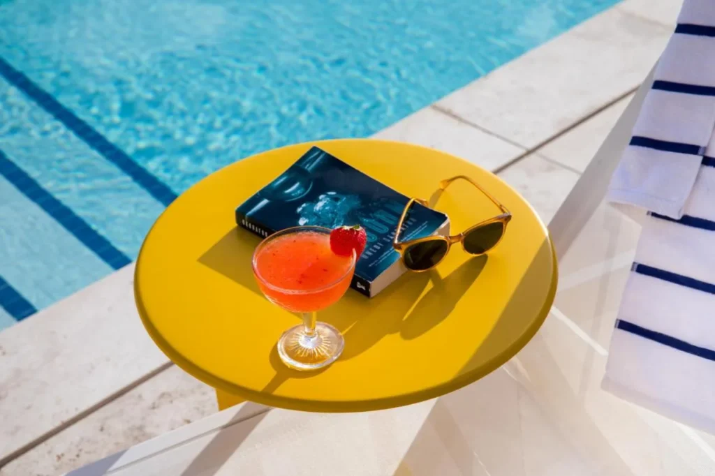 Cocktail, book and sunglasses on a yellow table at the pool edge, The Alfond Inn