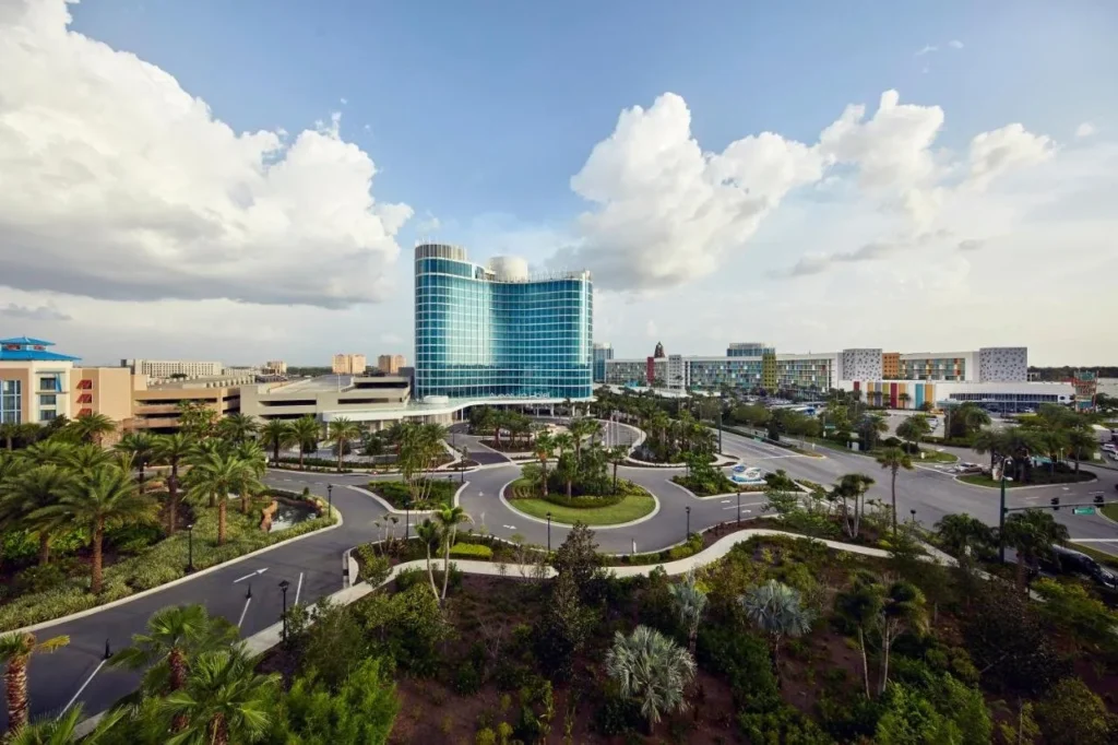 Vista elevada de la torre de vidrio curva y la entrada del Universal Aventura Hotel, con los coloridos edificios de Cabana Bay a la derecha