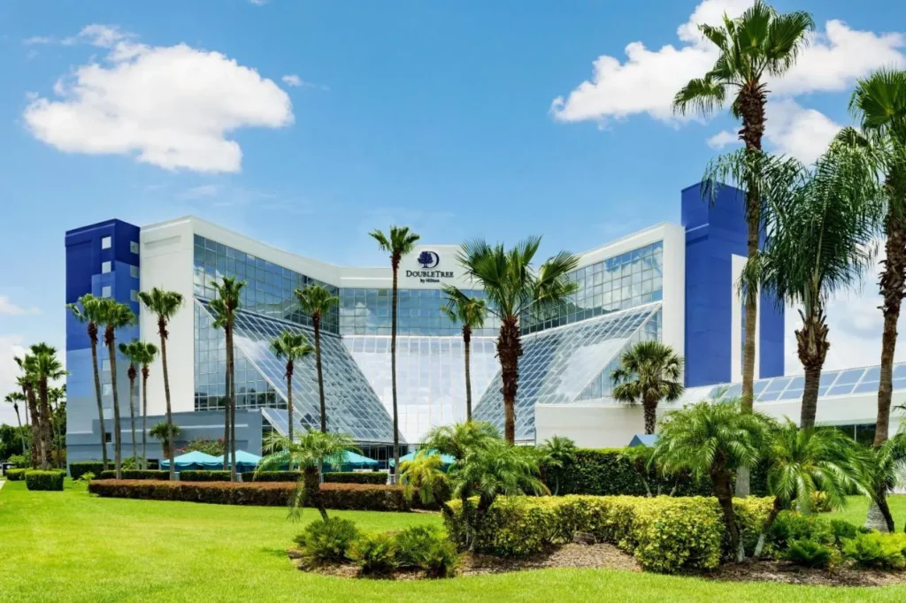 Edificio DoubleTree by Hilton Orlando Airport con fachada de vidrio detr&aacute;s de palmeras y un c&eacute;sped verde