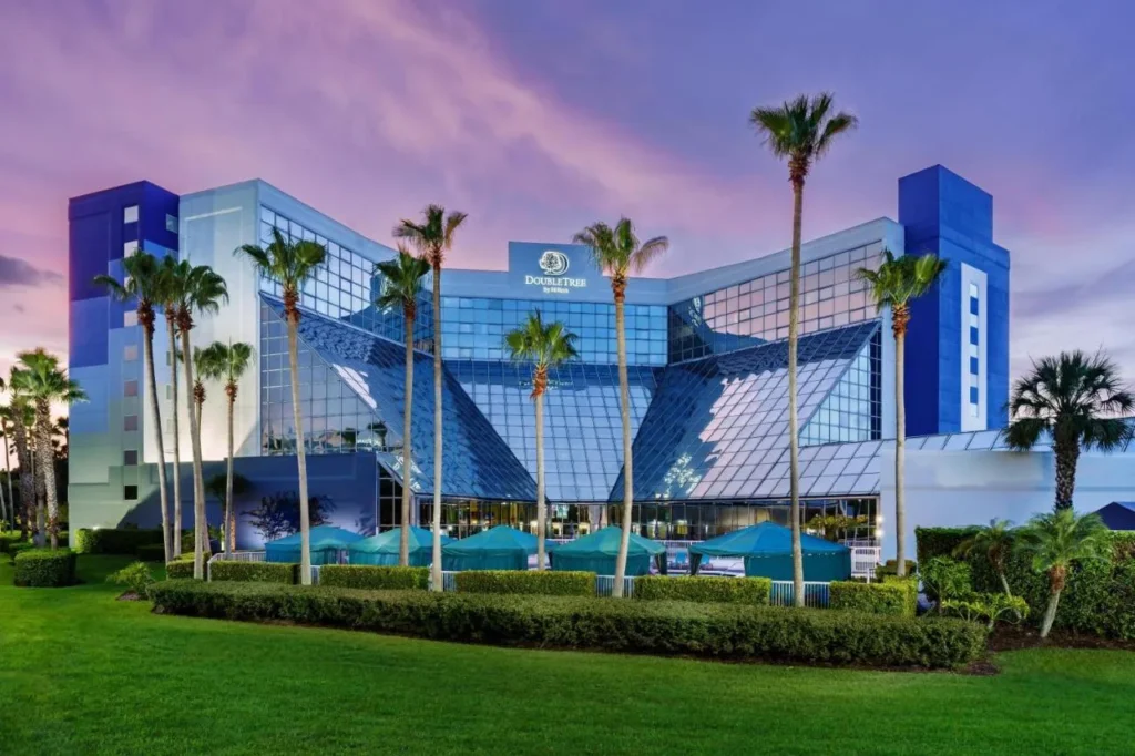 Glass-fronted DoubleTree by Hilton Orlando Airport building with palm trees and pool cabanas at dusk