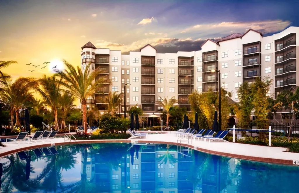 Torre de invitados con balcones detrás de una piscina bordeada de palmeras al atardecer en The Grove Resort