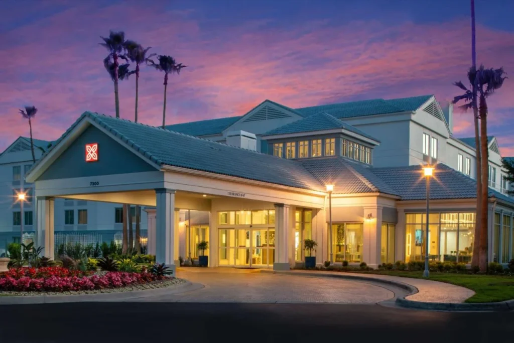 Hilton Garden Inn Orlando Airport dosel de entrada y vest&iacute;bulo iluminado al atardecer bajo las palmeras