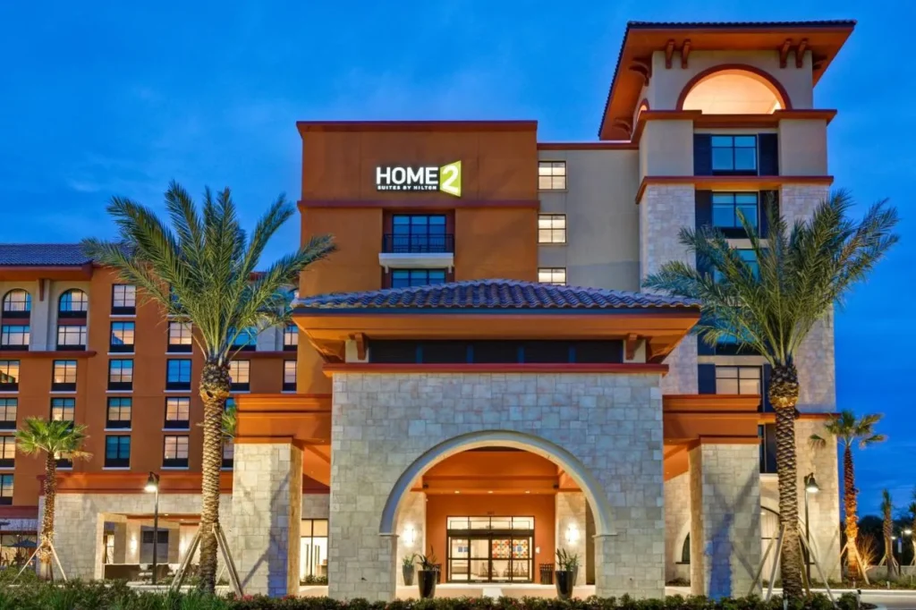 Torre Home2 Suites Orlando Flamingo Crossings y entrada de piedra arqueada al atardecer con palmeras