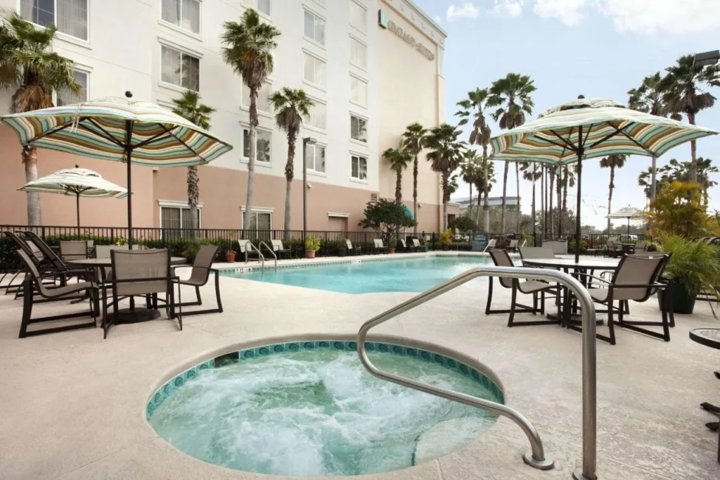Ba&ntilde;era de hidromasaje y piscina al aire libre con sombrillas junto a la torre del Embassy Suites Orlando Airport