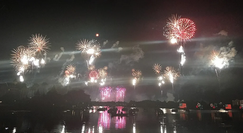 Independence Day fireworks above Magic Kingdom seen across a lagoon with boats and lit flag floats