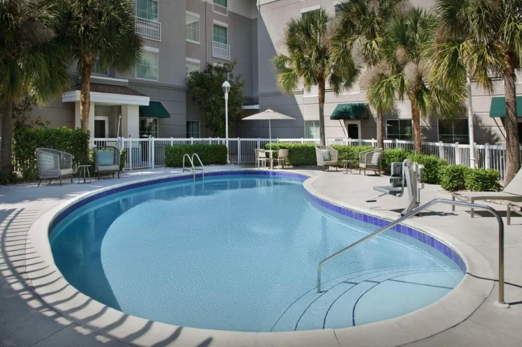 Piscina al aire libre en forma de ri&ntilde;&oacute;n con asientos en el patio y palmeras en Homewood Suites by Hilton Orlando Airport