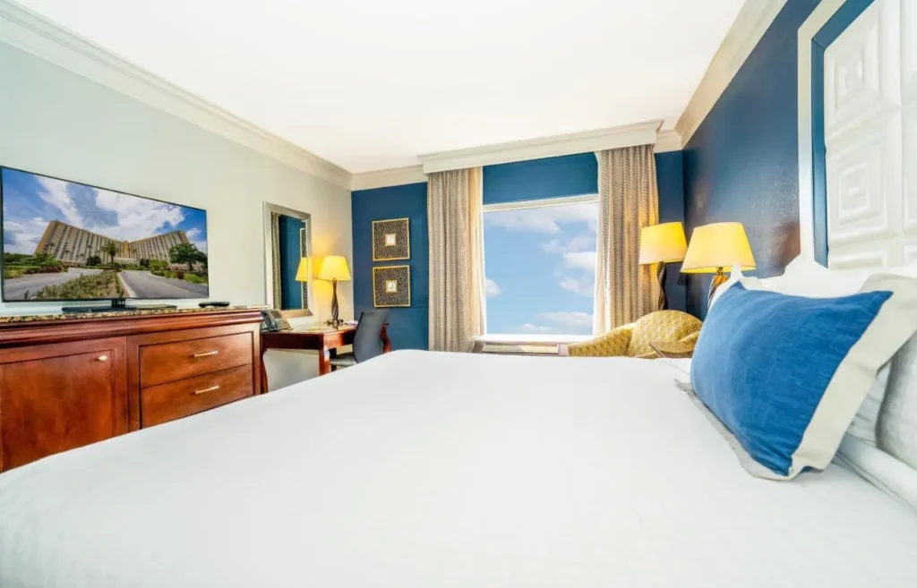 King bed facing a flat-screen TV, desk and bright window at Rosen Plaza Hotel