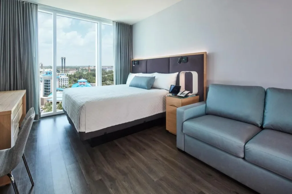 Cama king size, sof&aacute; gris-azul y escritorio de madera junto a una ventana alta en Universal Aventura Hotel