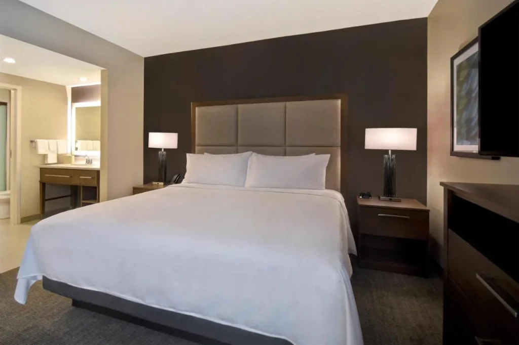 King bedroom with a lit vanity beyond in a Homewood Suites Flamingo Crossings suite