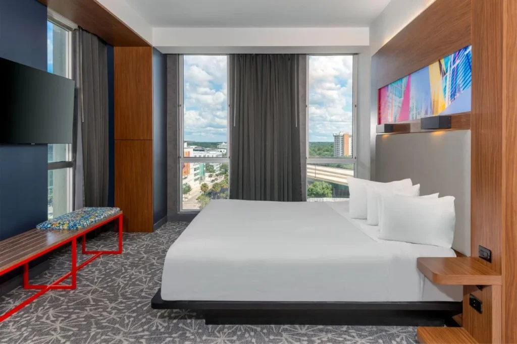 King room con un banco rojo, ventanas altas y vistas a la ciudad en el Aloft Orlando Downtown