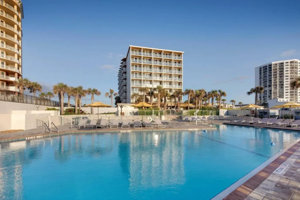 Large outdoor pool with loungers, palms and the hotel tower at Delta Hotels Daytona Beach Oceanfront