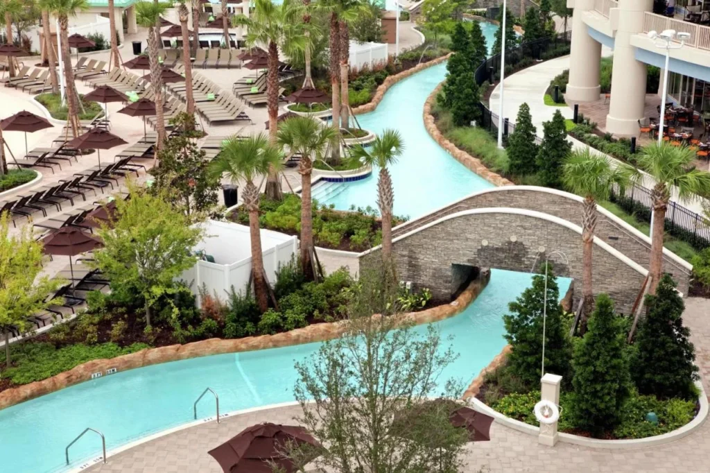 R&iacute;o perezoso que serpentea bajo un puente de piedra m&aacute;s all&aacute; de palmeras y tumbonas de la piscina en Signia by Hilton Orlando Bonnet Creek