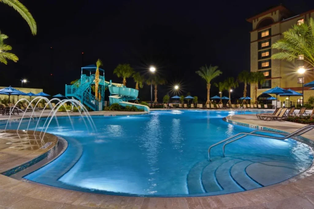 Lit pool with fountain jets and two waterslides at night at Home2 Suites Orlando Flamingo Crossings