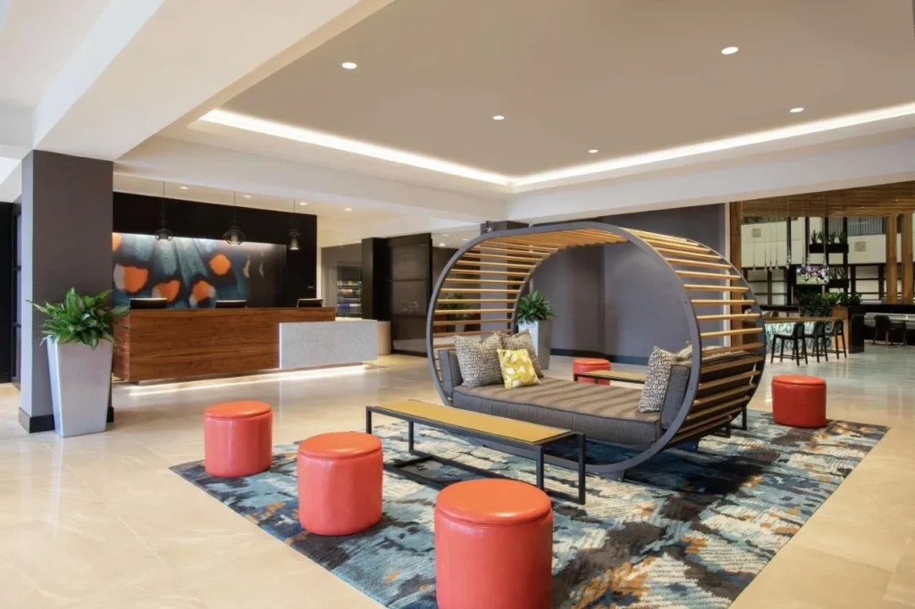 Lobby con un asiento de madera, otomanas naranjas y recepci&oacute;n en el Embassy Suites Orlando Airport