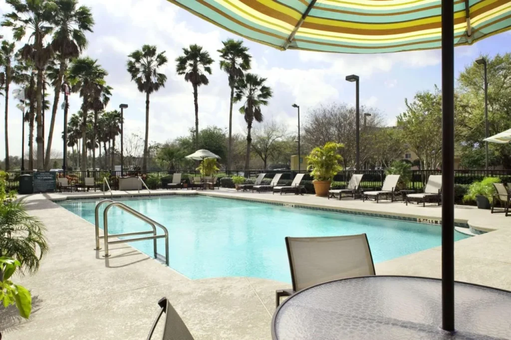 Outdoor pool with loungers, palm trees and striped umbrella at Embassy Suites by Hilton Orlando Airport