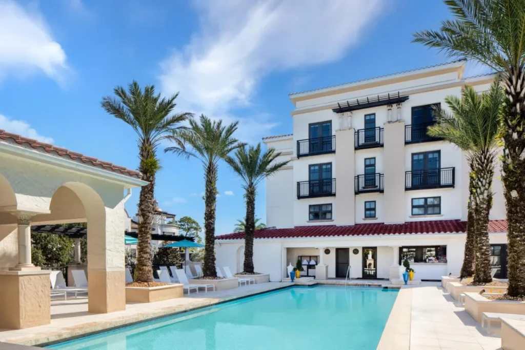 Piscina al aire libre con tumbonas, palmeras y un edificio blanco de estilo mediterr&aacute;neo con balcones en The Alfond Inn