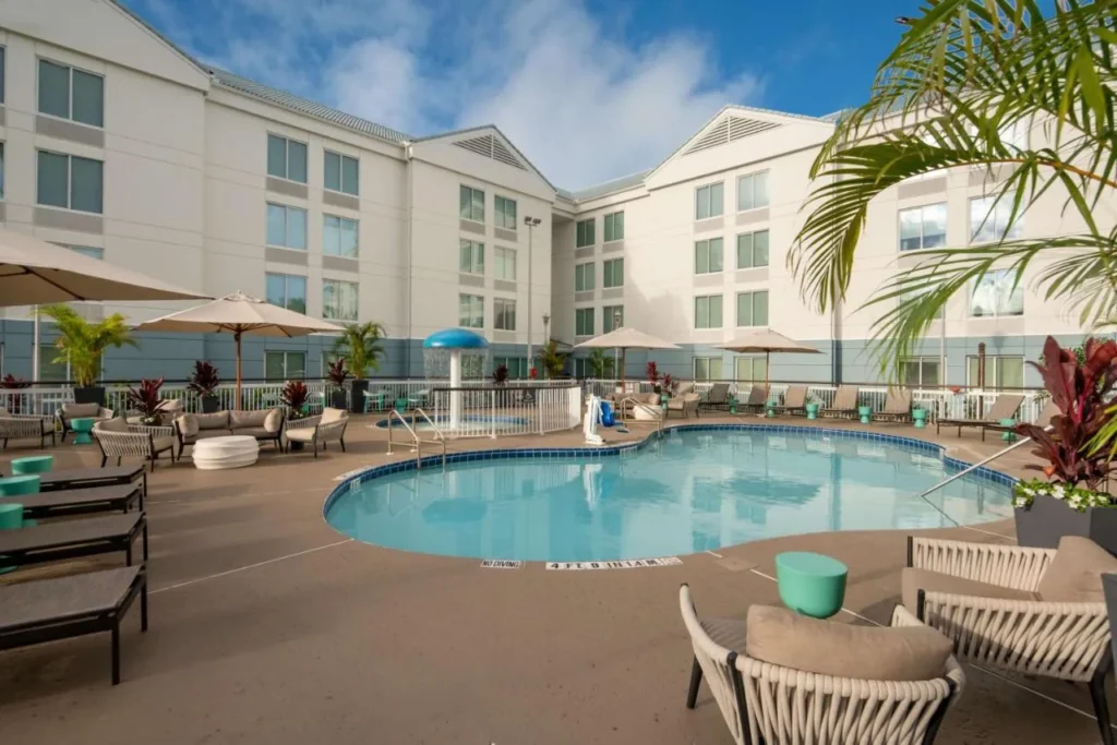 Piscina al aire libre con tumbonas, sombrillas y una fuente de hongos en Hilton Garden Inn Orlando Airport