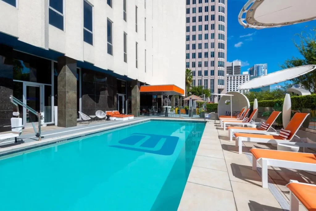 Piscina al aire libre con tumbonas naranjas junto al edificio blanco Aloft Orlando Downtown en un d&iacute;a soleado