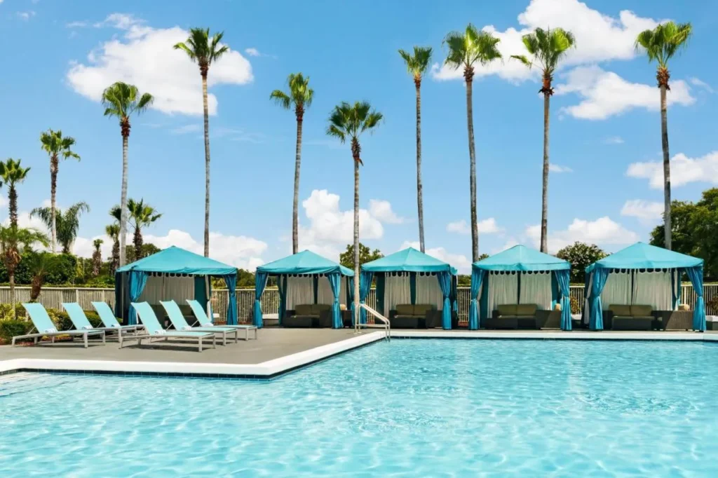 Piscina al aire libre con caba&ntilde;as turquesas y tumbonas bajo altas palmeras en DoubleTree Orlando Airport