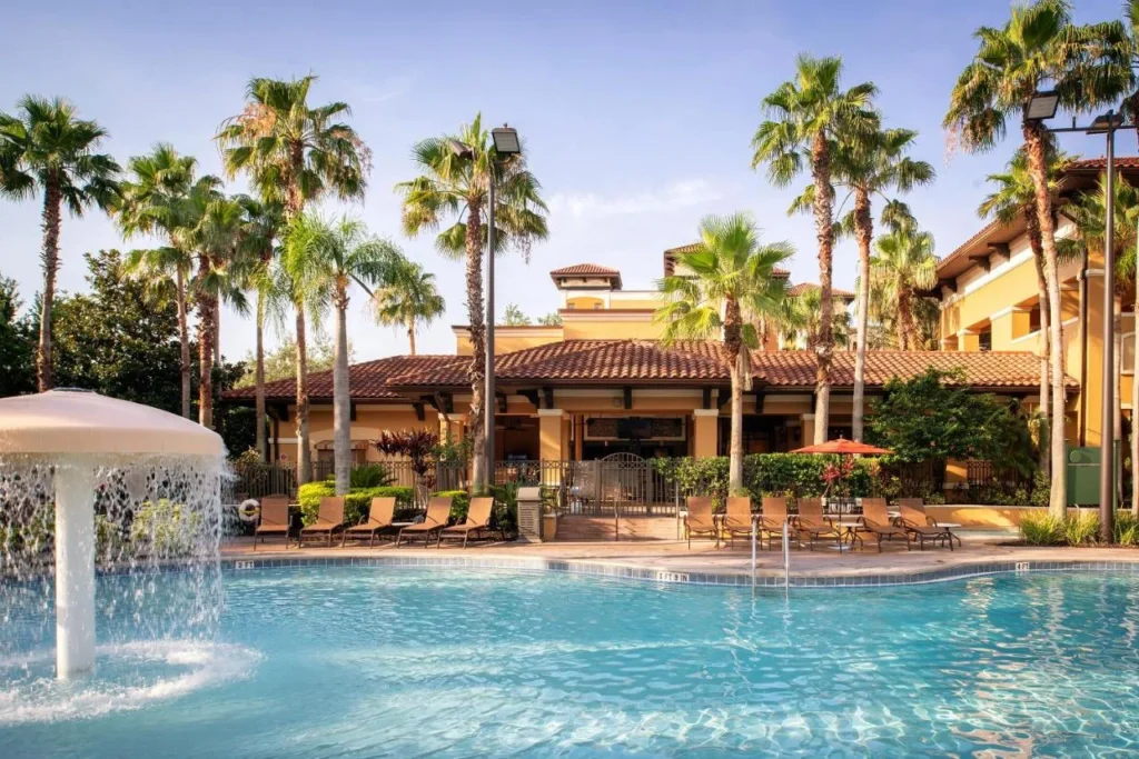 Piscina bordeada de palmeras con una fuente de hongos frente a la casa club en Floridays Resort Orlando