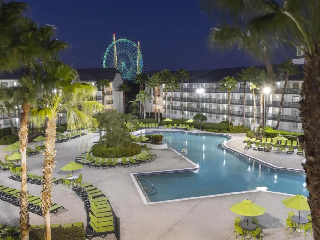 Piscina y tumbonas verdes por la noche con el Orlando Eye iluminado detrás de Avanti International Resort