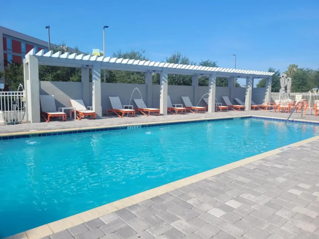 Piscina exterior rectangular con tumbonas naranjas bajo una p&eacute;rgola blanca en el Holiday Inn & Suites Orlando I-Drive Theme Parks
