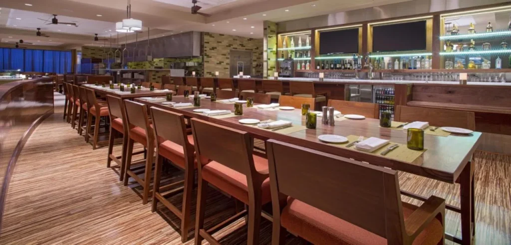 Restaurante con mesas largas de madera, sillas anaranjadas y un bar retroiluminado en el Hyatt Regency Orlando Airport