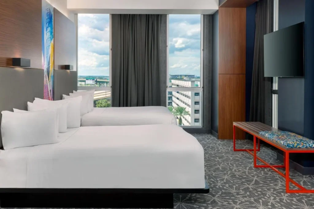Room with two beds, a red bench and tall windows over downtown at Aloft Orlando Downtown
