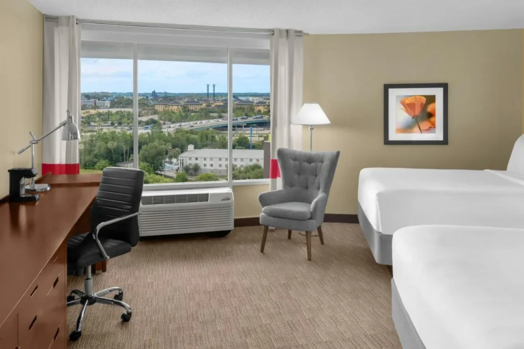 Habitación con dos camas, escritorio y una gran ventana con vistas a la ciudad en Four Points I-Drive