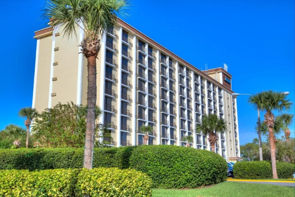 Rosen Inn M&aacute;s cercano a la torre Universal detr&aacute;s de palmeras y setos recortados en un d&iacute;a soleado