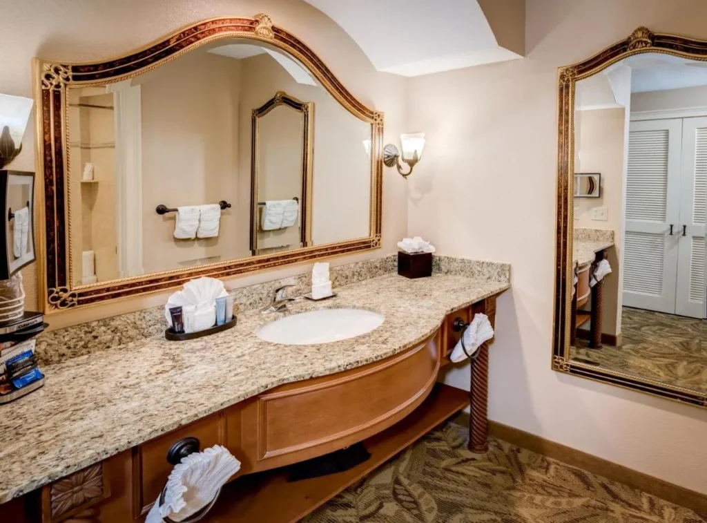 Rosen Shingle Creek room vanity with a granite counter, gold-framed mirrors and fanned towels