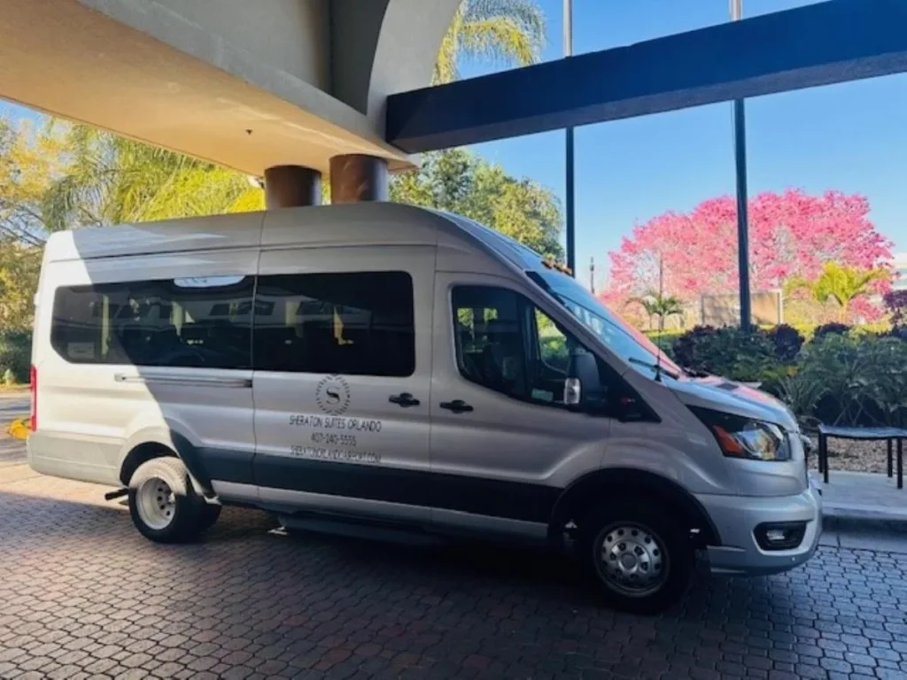 Sheraton Suites Orlando Airport shuttle van parked under the entrance canopy beside flowering trees