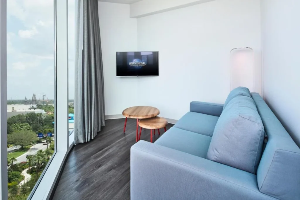 &Aacute;rea de descanso con sof&aacute; azul en Universal Aventura Hotel, junto a una ventana de piso a techo que mira hacia los parques tem&aacute;ticos