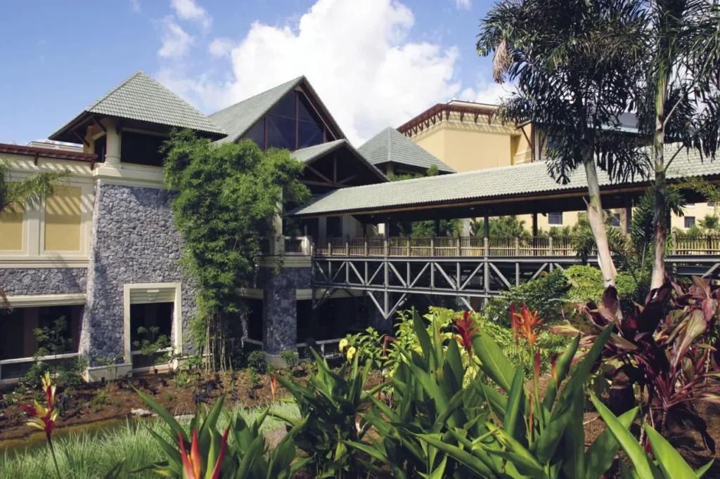 Edificio de vest&iacute;bulo revestido de piedra y pasarela cubierta en medio de plantas tropicales en Loews Royal Pacific Resort