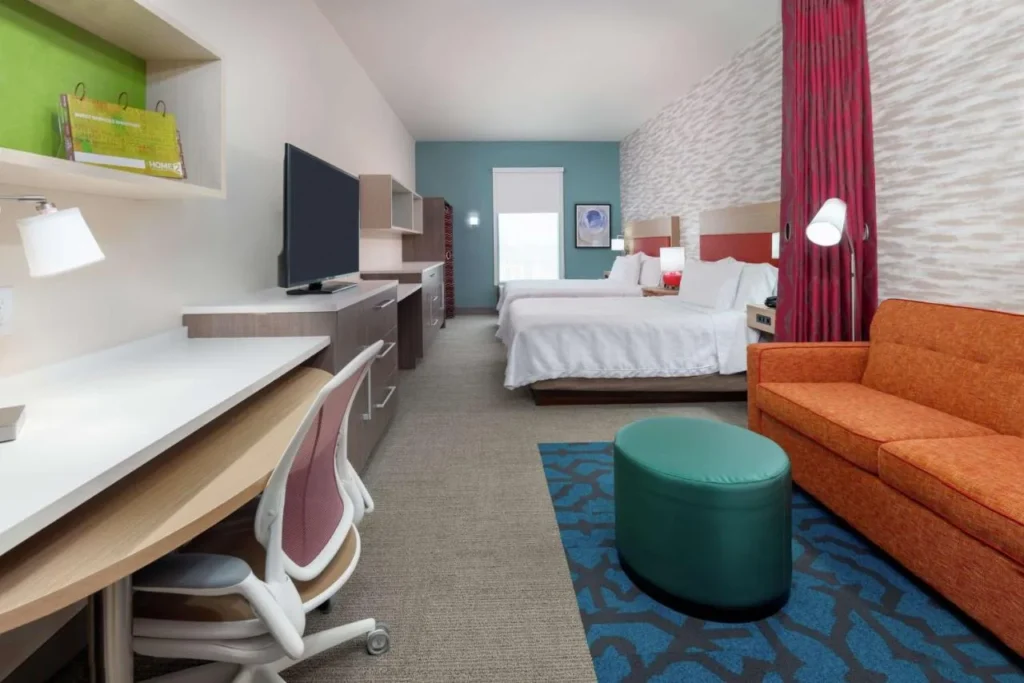 Suite tipo estudio con dos camas queen, sofá naranja y escritorio largo en Home2 Suites Near Universal