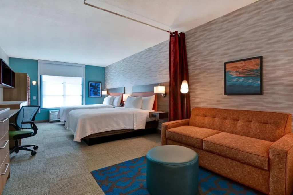 Suite estudio con dos camas queen, sofá cama naranja y escritorio en Home2 Suites Flamingo Crossings
