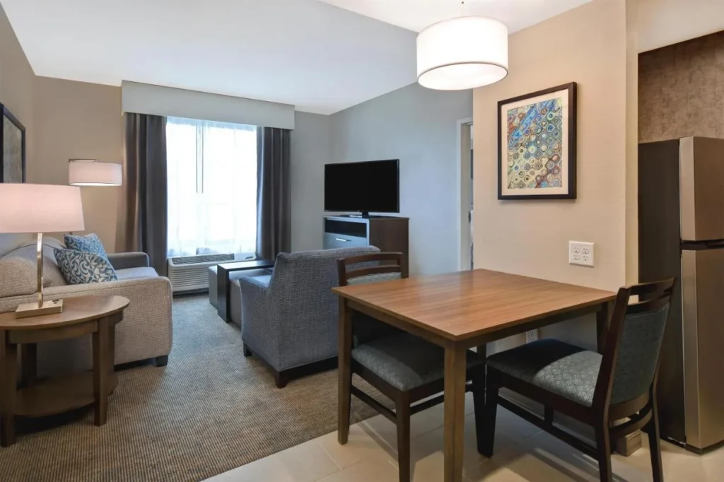 Suite living room with sofa, armchair, TV, dining table and fridge at Homewood Suites Flamingo Crossings