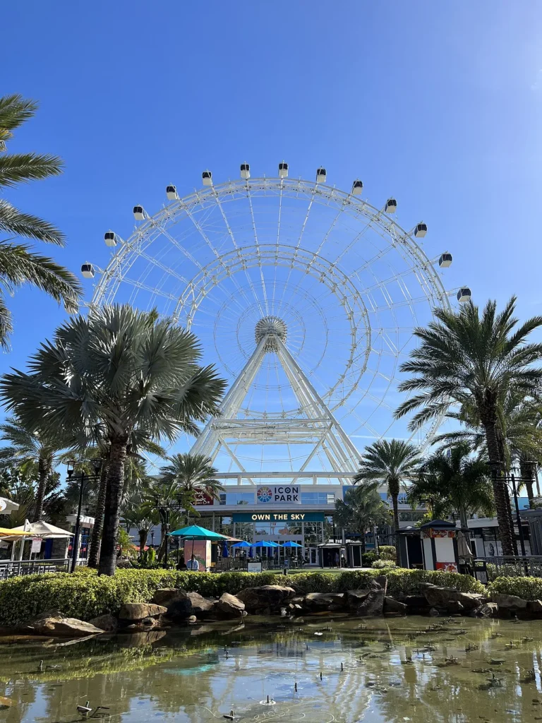 The Orlando Eye observation wheel rising above palm trees and a pond at ICON Park Orlando
