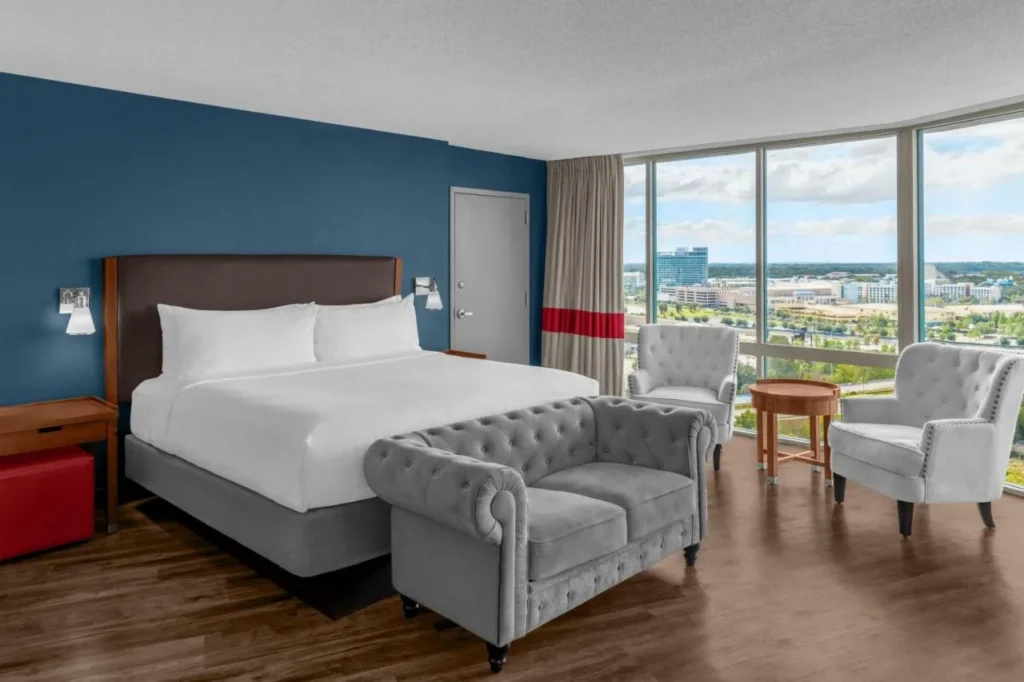 Habitación en planta alta con cama grande, sofá cama gris y pared de ventanas en Four Points I-Drive
