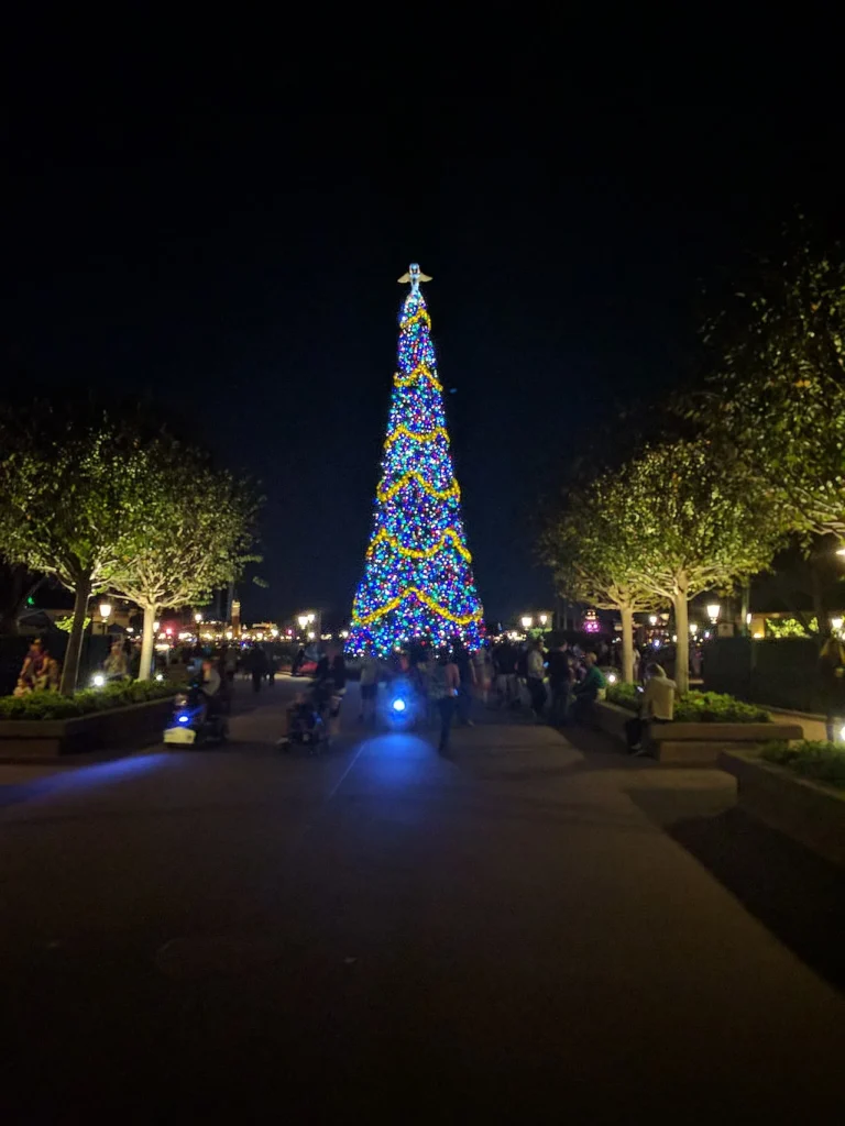 Towering Christmas tree glowing with colorful lights at the end of an EPCOT walkway at night