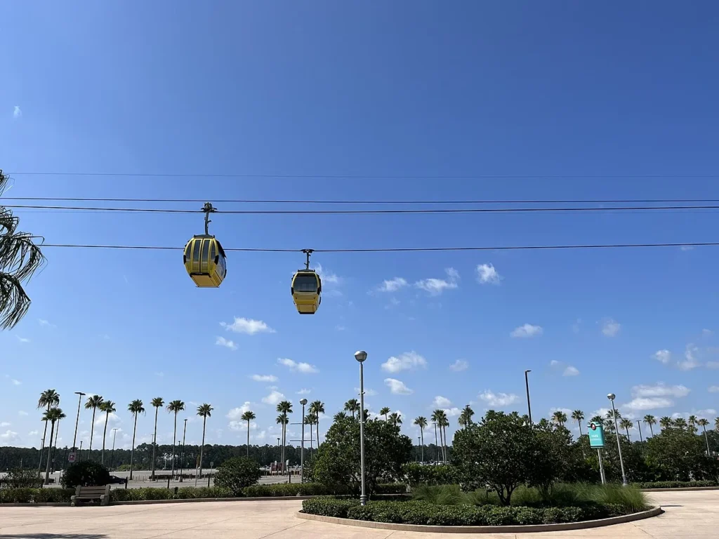 Two yellow Disney Skyliner gondolas on their cables above palm trees and a theme park plaza