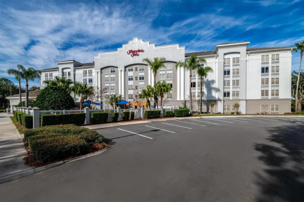 Edificio blanco de cinco pisos Hampton Inn Orlando/Lake Buena Vista con palmeras y su estacionamiento