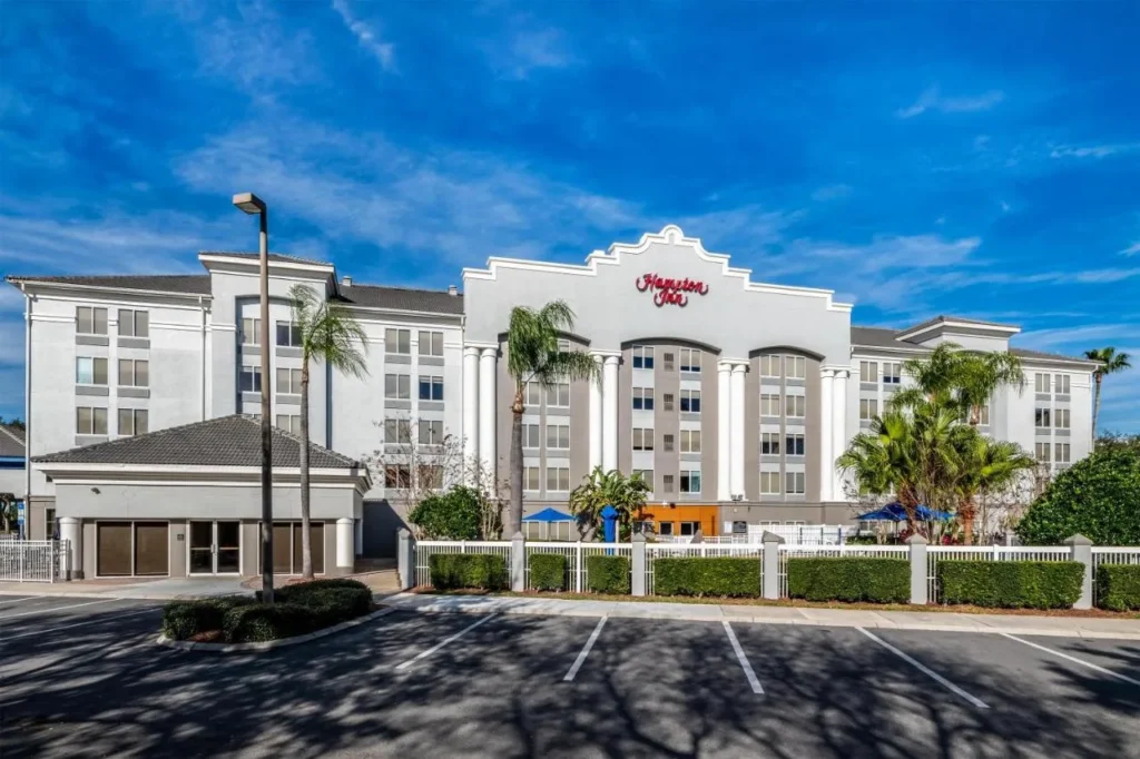 Edificio hotelero blanco con palmeras y estacionamiento en Hampton Inn Orlando/Lake Buena Vista