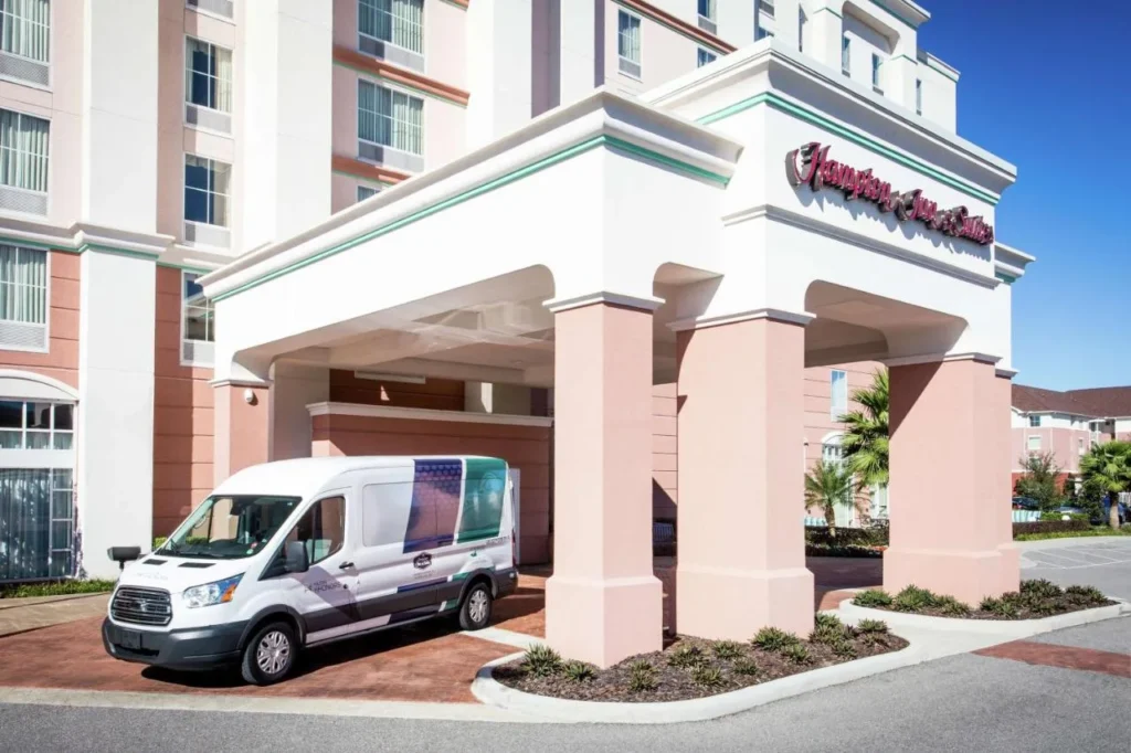 White shuttle van under the entrance canopy of Hampton Inn & Suites Orlando Airport Gateway Village